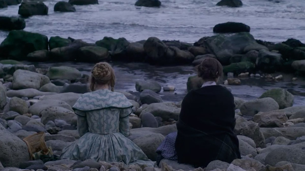 Two women sit with their backs to the camera facing the sea in a still from Ammonite.